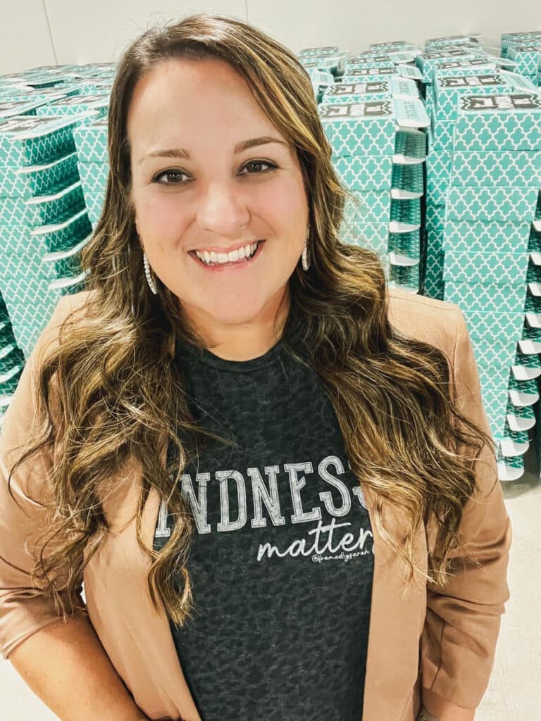Sarah Williams in black leopard print _Kindness Matters_ Tshirt and Camel blazer standing in front of a stack of teal Monogram Boxes