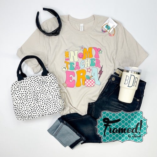 Cream "In My teacher Era" graphic tshirt styled with dark wash jeans, monogrammed tumbler, black and white spotted lunch bag, colorful earrings, and black headband.