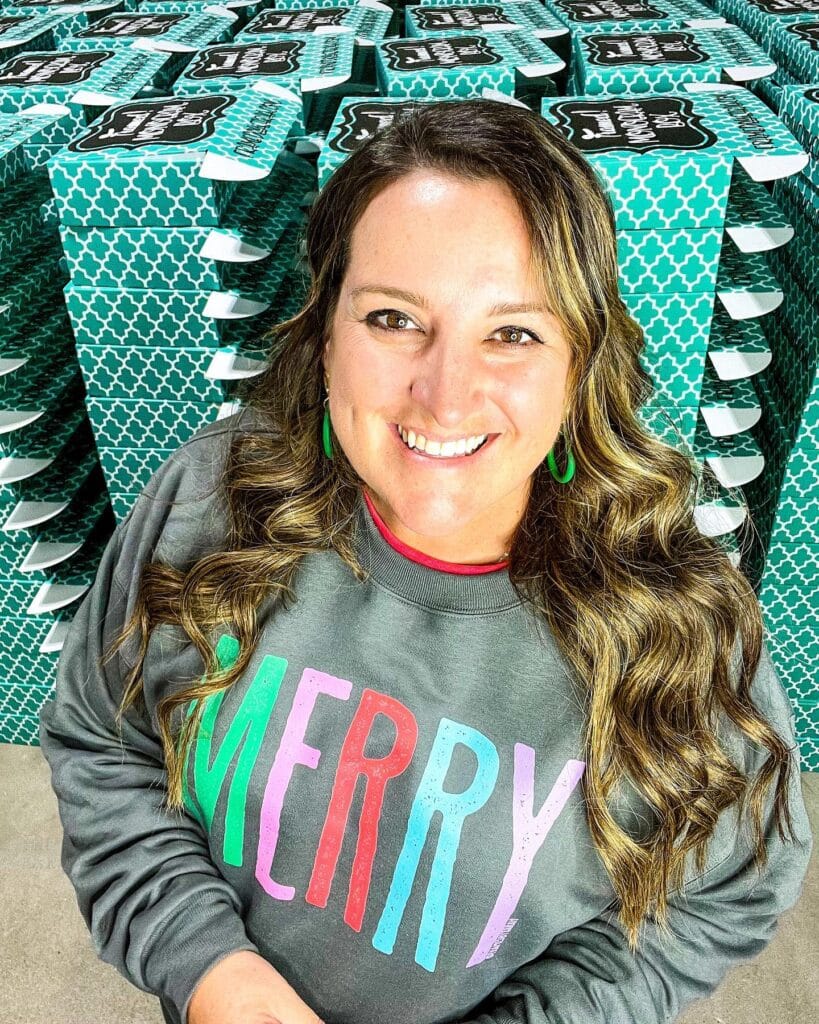 Sarah Williams standing in front of Monogram Box stack wearing a gray "Merry" Holiday sweatshirt