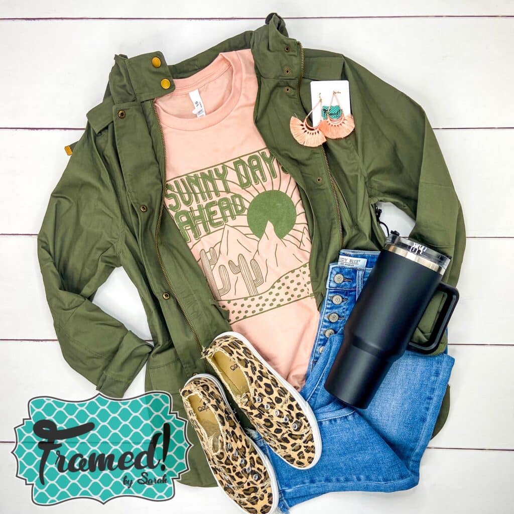 Peach shirt with a desert scene and the word Sunny Days Ahead in olive green ink. Styled with Jeans and olive utility jacket