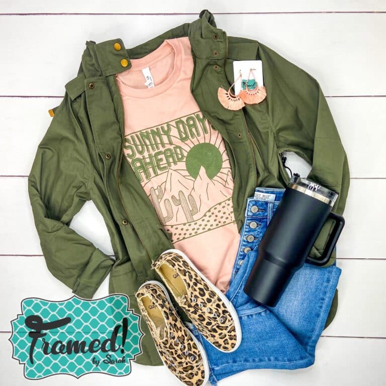Peach shirt with a desert scene and the word Sunny Days Ahead in olive green ink. Styled with Jeans and olive utility jacket