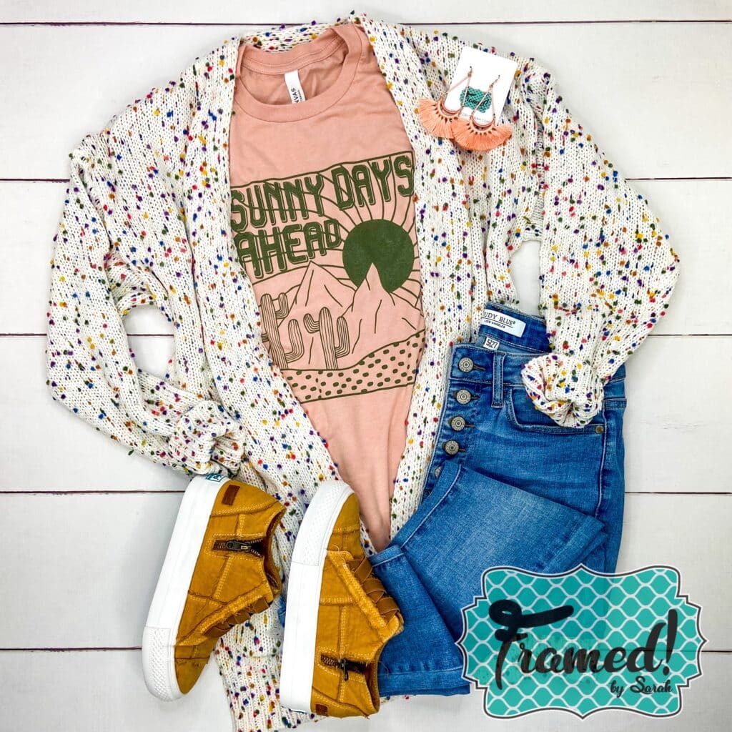 Peach shirt with a desert scene and the word Sunny Days Ahead in olive green ink. Styled with Jeans and camel shoes and white confetti cardigan