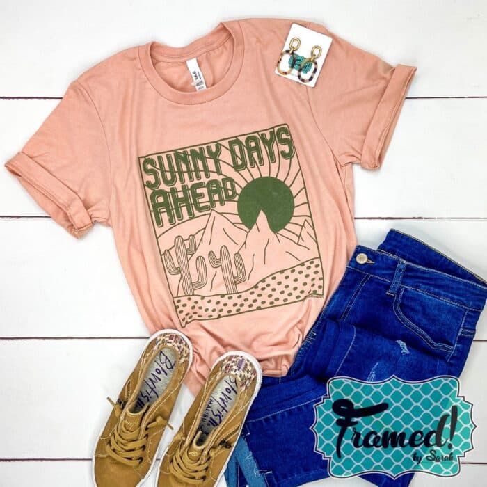 Peach shirt with a desert scene and the word Sunny Days Ahead in olive green ink. Styled with Jeans and camel shoes