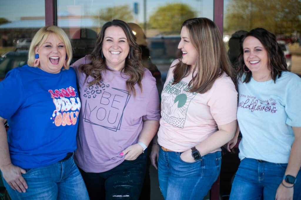 4 Women in custom designed graphic tees standing in front of a building with large window