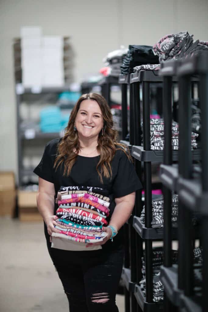 Sarah Williams in a black shirt, standing in front of rows of shelves, holding a stack of folded colorful t-shirts