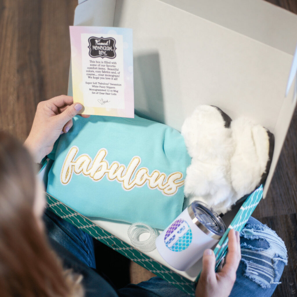 over the shoulder look at a woman opening a Monogram Box. The box is full of a teal "Fabulous" Sweatshirt, white fluffy slippers, a monogrammed tumbler, and hair ties.