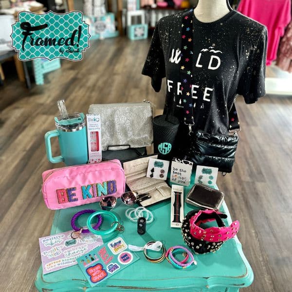 table full of fun items perfect for a teen girl summer survival kit. Items include: Black Wild and Free graphic tee, large turquoise tumbler with handle, black studded tumbler with monogram, pink Be Kind zipper pouch, jeweled headbands, stickers, bracelets, wallet, puffer crossbody purse, and more.