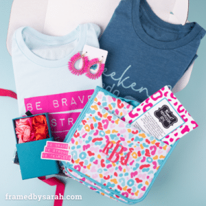 March 2024 Monogram contents displayed in an open box. Contents include: Navy graphic tee, Light blue and hot pink graphic tee, colorful leopard print travel bag with hot pink monogram, hot pink drop hoop earrings, small blue box holding 3 silk scrunchies. in shades of pink and red