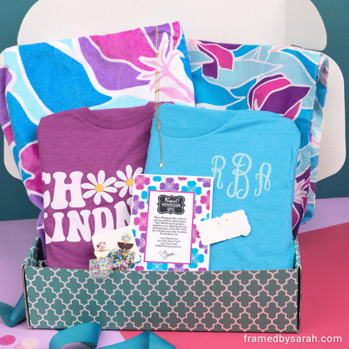 May 2025_Monogram Box and T-Shirt Club displayed inside an open turquoise box and bag. Contents include: blue monogrammed t-shirt, floral print towel, matching floral print kimono, gold ankle bracelet, colorful acrylic earrings, "choose kindness" purple shirt