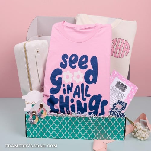 Pink See Good In All Things T-shirt, Ombre Raffia wrapped earrings, white makeup bag, monogrammed sweatshirt, cream and gold dainty bracelet displayed in a teal subscription box