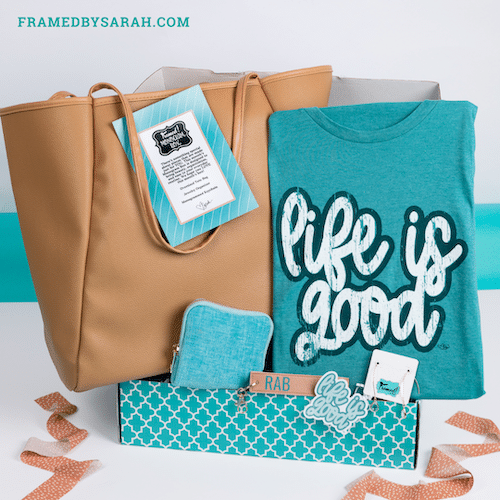 Blue "life is good" tshirt, oversized tote, monogrammed key fob, turquoise jewelry pouch, and crystal drop earrings displayed in a teal subscription box