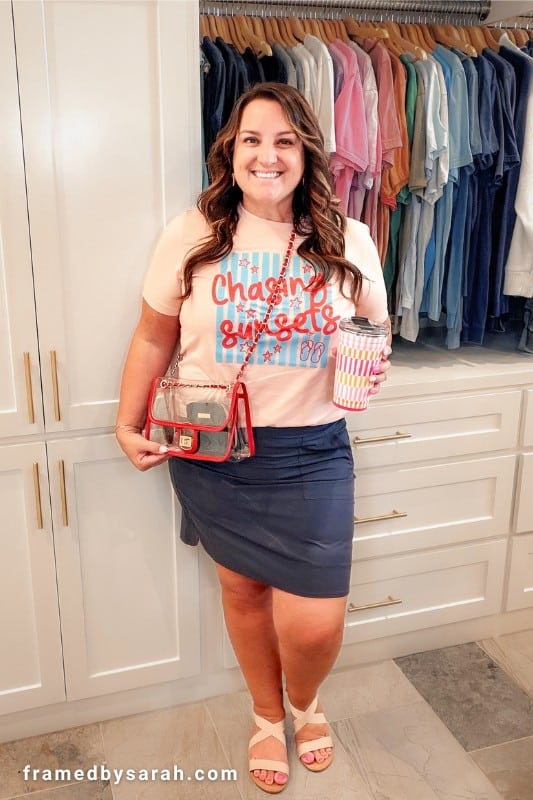 Sarah wearing the "chasing sunsets" tshirt styled with a navy skirt, sandals, red trim stadium bag, and tumbler
