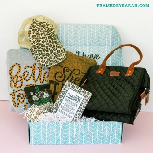 teal box open and displaying gray better together tee shirt, black quilted cooler tote bag, leopard print trucker hat, leopard earrings, and a bag of pretzels