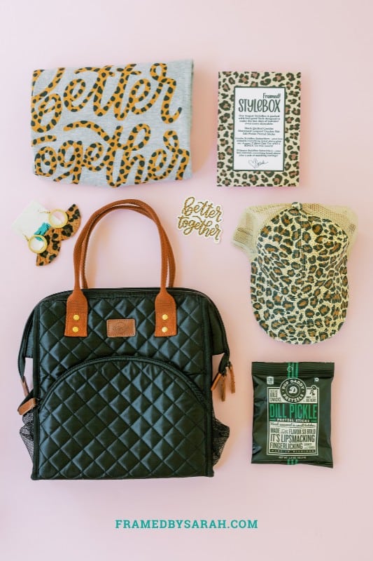 Pink background with gray better together tee shirt, black quilted cooler tote bag, leopard print trucker hat, leopard earrings, and a bag of pretzels on display