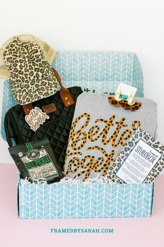 teal box open and displaying gray better together tee shirt, black quilted cooler tote bag, leopard print trucker hat, leopard earrings, and a bag of pretzels
