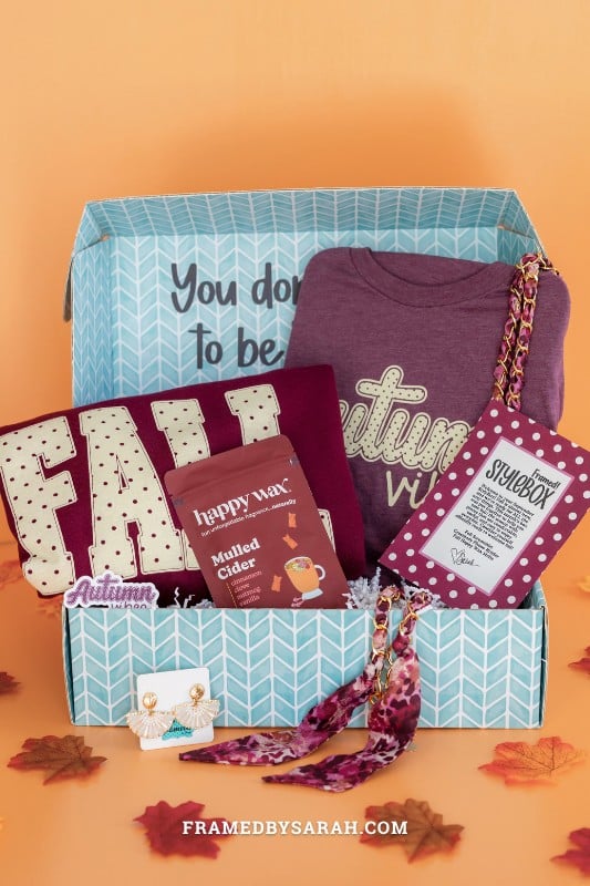teal Framed Style Box open displaying the contents which include, maroon "fall" sweatshirt, scarf phone chain, cider wax melts, and print insert.
