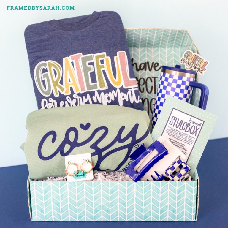 October 2026 Ultimate StyleBox contents displayed inside the teal open box. Content includes: seafoam "cozy season" sweatshirt, navy "grateful for every moment" tee, blue and white checkered tumbler, blue and white hair clip set, colorful beaded earrings, and sticker