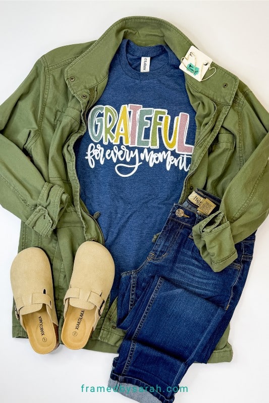 Flatlay photo of Navy "Grateful for Every Moment" t-shirt styled with green jacket, slide shoes, dark wash jeans, colorful earrings