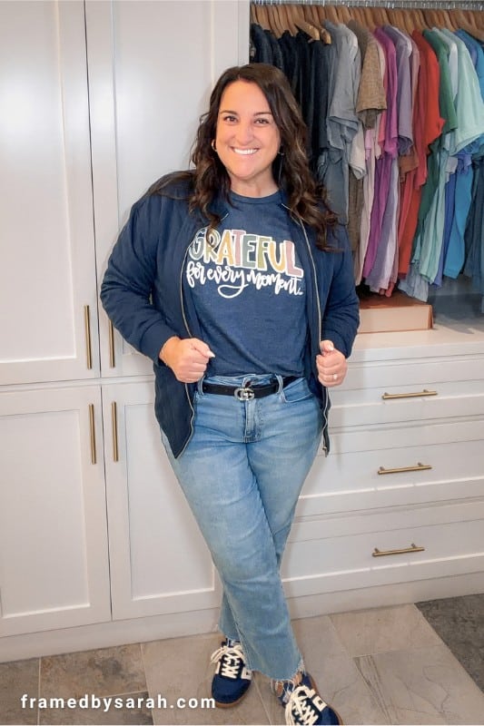 Sarah Williams wearing the navy "Grateful for Every Moment" t-shirt styled with jeans, navy jacket, navy sneakers, and colorful earrings